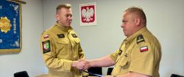 Komendant Powiatowy Państwowej Straży Pożarnej wręcza nagrodę funkcjonariuszowi podczas uroczystego spotkania służbowego. Obaj strażacy w umundurowaniu ściskają sobie dłonie. W tle widoczne są godło Rzeczypospolitej Polskiej oraz sala konferencyjna Komendy Powiatowej PSP.
