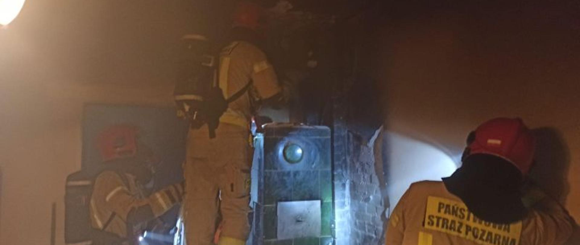 Zdjęcie wykonane wewnątrz pomieszczenia w którym wybuchł pożar. Strażacy stoją przy piecu kaflowym, jest lekkie zadymienie. Strażacy mają na sobie piaskowe mundury, hełmy czerwone i aparaty ochrony dróg oddechowych.