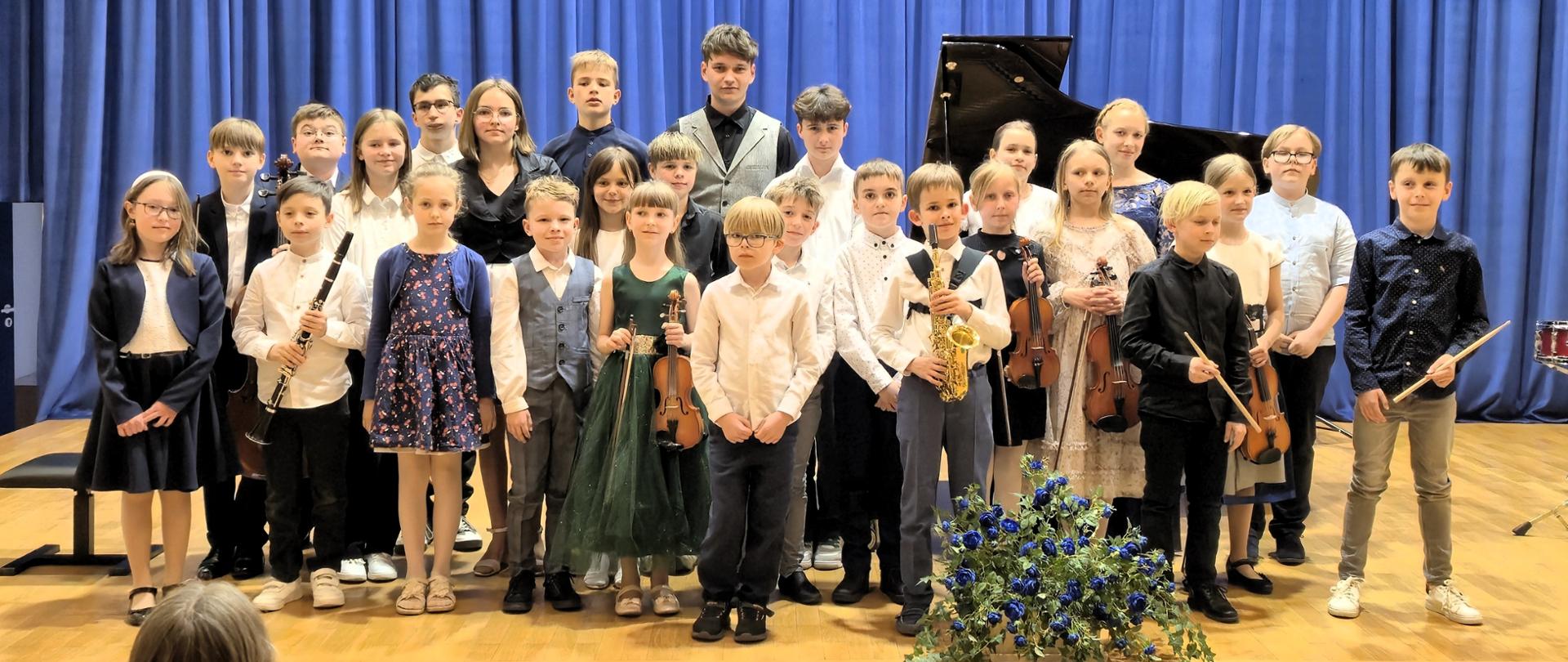 grupa kilkudziesięciu dzieci na scenie ustawionych w kilu rzędach, niektórzy trzymają instrumenty, przed nimi bukiet niebieskich kwiatów, w tle niebieska kotara