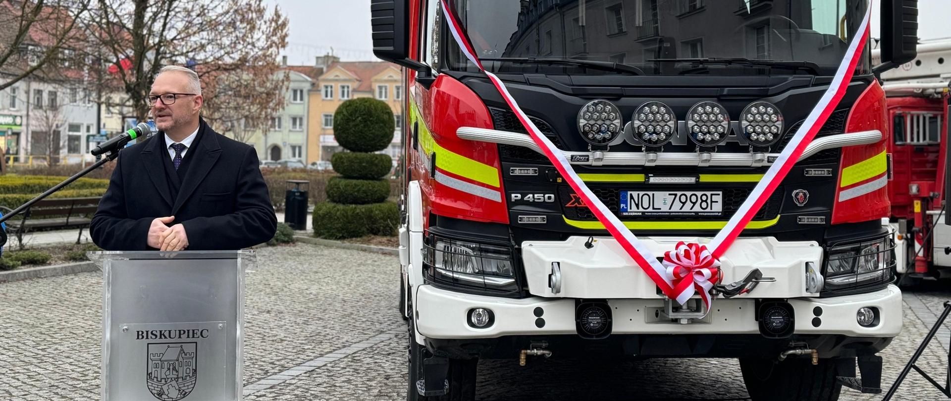 Mężczyzna przemawia przed wozem strażackim 