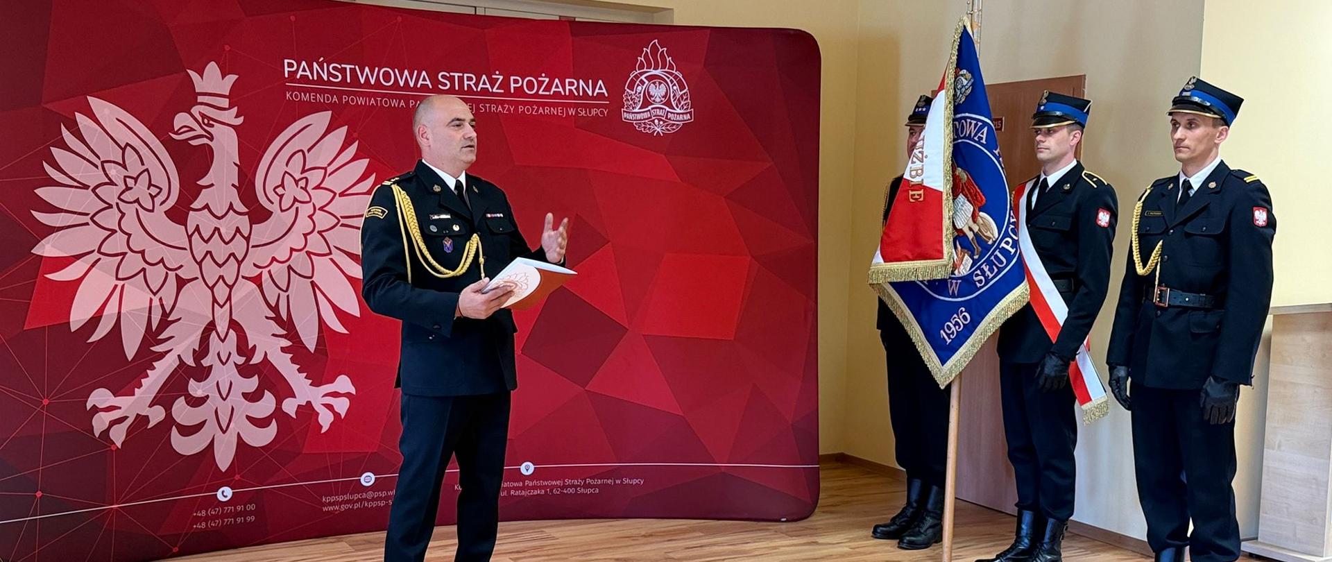 Komendant przemawia na tle ścianki z boku poczet sztandarowy 