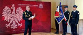 Komendant przemawia na tle ścianki z boku poczet sztandarowy