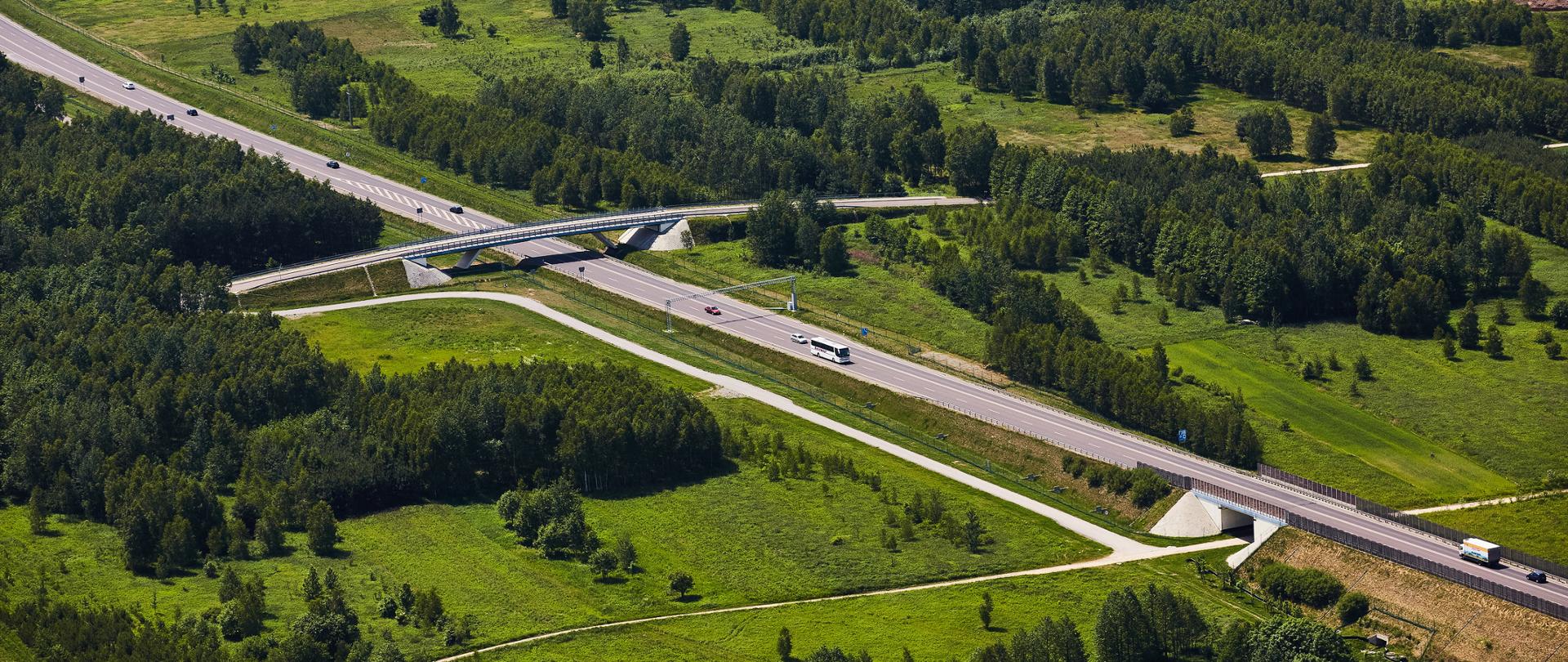 Zdjęcie przedstawia widok z lotu ptaka na odcinek drogi szybkiego ruchu przebiegający przez zielony, zalesiony teren. W centralnej części widać estakadę lub wiadukt przecinający trasę, otoczony gęstą roślinnością. Droga biegnie przez szerokie, otwarte przestrzenie z polami, łąkami i pasami lasu, a po jezdni poruszają się pojedyncze pojazdy. Całość tworzy harmonijny krajobraz infrastruktury drogowej wkomponowanej w naturalne otoczenie.
