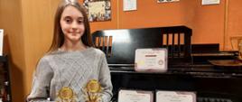 Zdjęcie kolorowe. Dziewczynka, blondynka w popielatym sweterku siedzi przy fortepianie, w rękach trzyma złote trofea - nagrody z konkursu w Londynie, na pulpicie i klawiaturze fortepianu stoją dyplomy, które otrzymała Alicja.