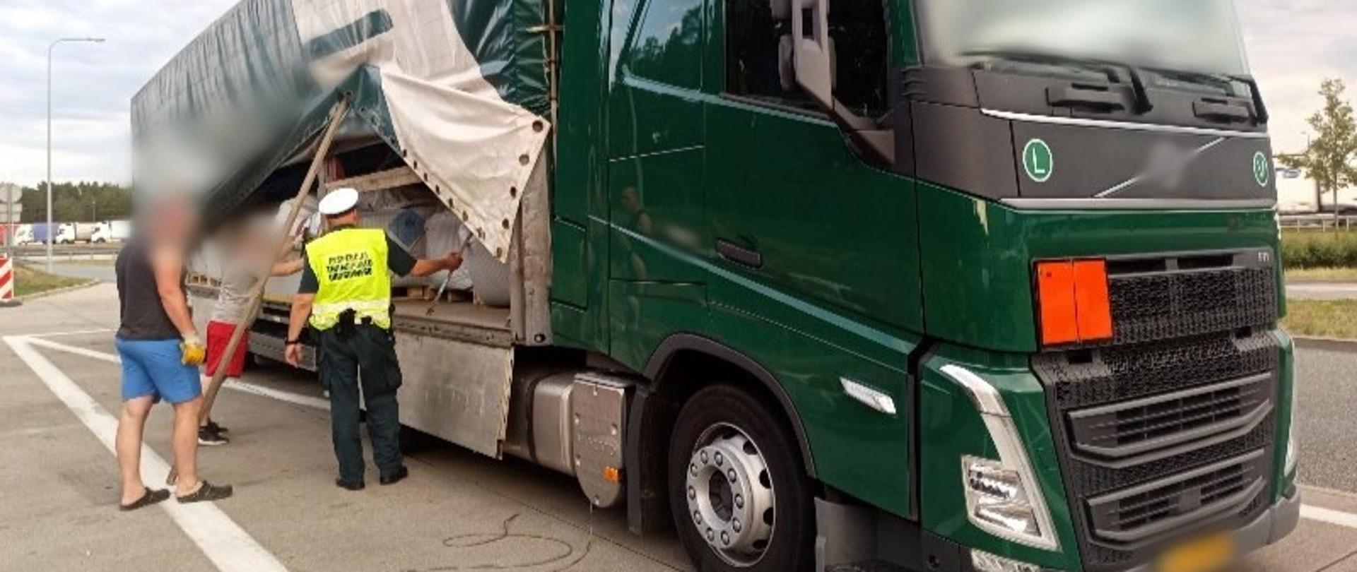 Zatrzymany do kontroli drogowej zestaw ciężarowy oznaczony pomarańczową tablicą z przodu (ADR). Inspektor stoi obok plandeki podniesionej na naczepie przez kierowców ciężarówki i kontroluje, co jest przewożone w ładowni. 