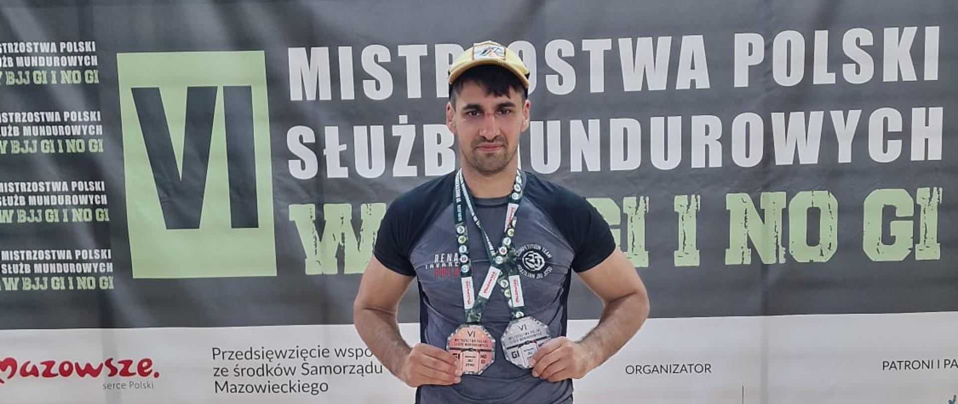 Strażak w stroju sportowym stoi przed podium i prezentuje dwa medale zdobyte podczas VI Mistrzostw Polski Służb Mundurowych w Brazylijskim Jiu-Jitsu Gi i No Gi.