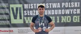 Strażak w stroju sportowym stoi przed podium i prezentuje dwa medale zdobyte podczas VI Mistrzostw Polski Służb Mundurowych w Brazylijskim Jiu-Jitsu Gi i No Gi.