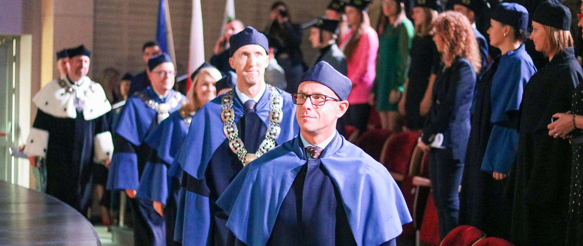 Ludzie w niebieskich ceremonialnych strojach idą rzędem przez salę, z boku widać krzesła.