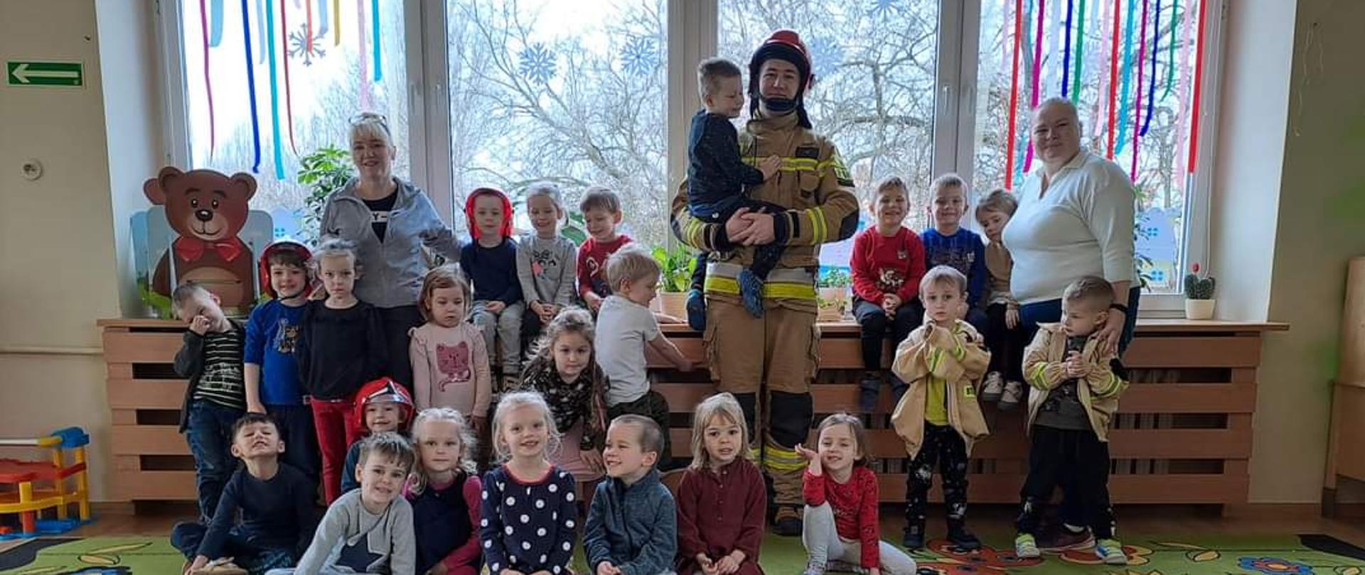 Na zdjęciu widzimy strażaka ubranego w odzież ochronną koloru piaskowego na głowie czerwony hełm. Strażak pozuje razem z dziećmi do zdjęcia grupowego.