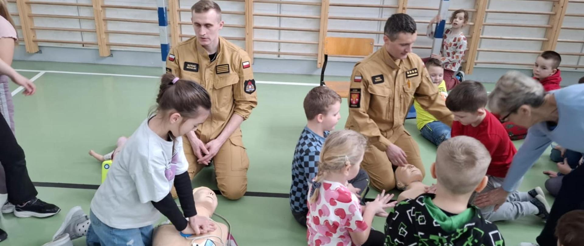 na podłodze klęczą dwaj strażacy a naokoło nich dzieci ćwiczą udzielanie pierwszej pomocy