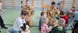 na podłodze klęczą dwaj strażacy a naokoło nich dzieci ćwiczą udzielanie pierwszej pomocy