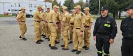 Strażacy podczas przywitania z Zastępcą Komendanta Głównego oraz Komendantem Wojewódzkim. Ustawieni w dwuszeregu stoją na placu ubrani w piaskowe mundury oraz czarne ubrania koszarowe. Komendanci witają się z załogą poprzez podanie ręki z tyłu widoczne budynki oraz drzewa