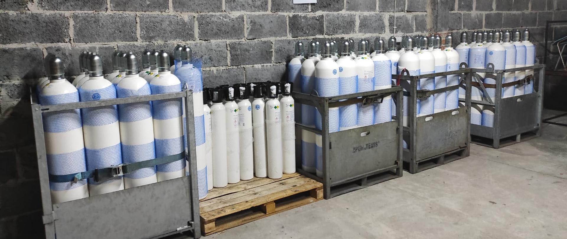 Na zdjęciu widać składowane butle z tlenem w metalowych pojemnikach ora na drewnianej palecie. Za butlami widać mur z szarego pustaka. Metalowe skrzynie i paleta są rozmieszczone na betonowej posadzce. Nad butlami wisi kartka papieru A4.
