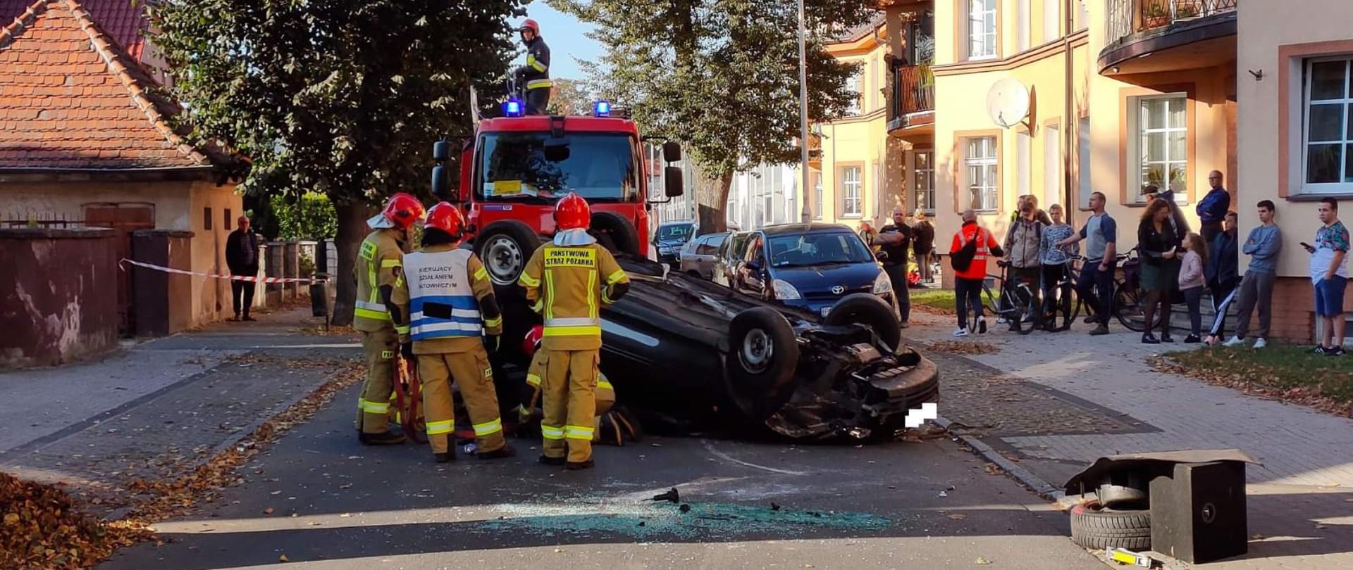 Strażacy przy rozbitym aucie. Wokół dużo ludzi.