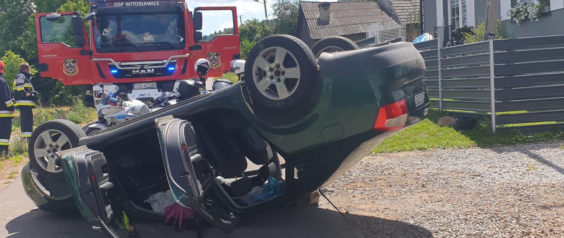 Na zdjęciu widać służby ratownicze oraz wywrócony do góry kołami na jezdni samochód osobowy marki AUDI koloru zielonego. W tle zaparkowane dwa motocykle policyjne oraz samochód strażacki. Przy samochodzie gaśniczym stoi dwóch strażaków.