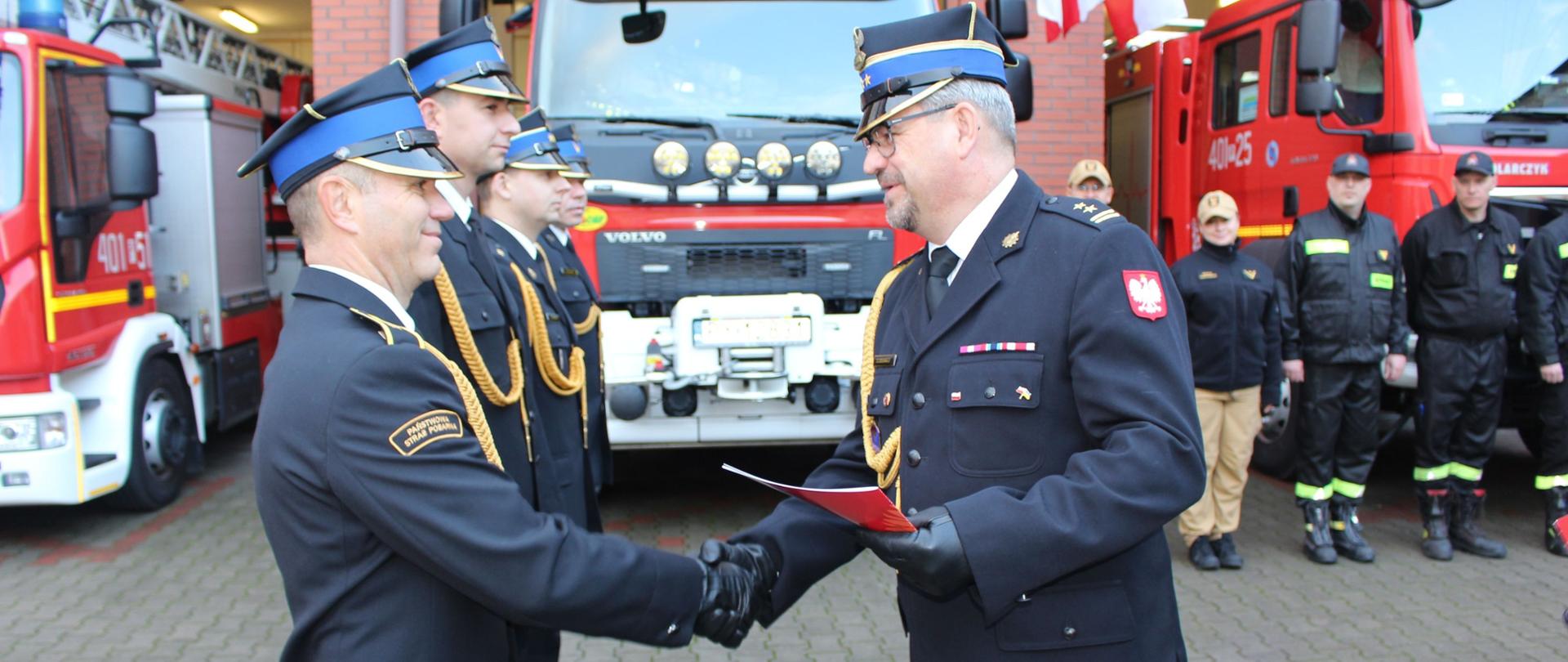 Zdjęcie przedstawia strażaków w trakcie uroczystego wręczania awansów.
W tle budynki.
