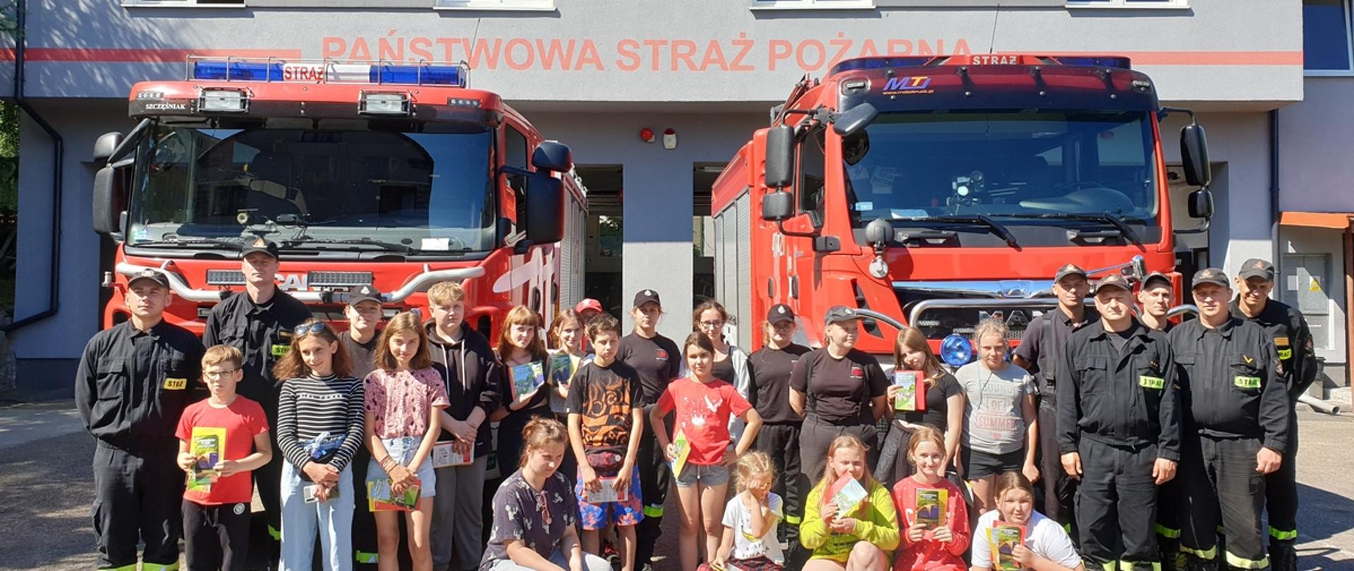 Dzieci ze strażakami przy dwóch samochodach strażackich, w tle strażnica Jednostki Ratowniczo gaśniczej w Braniewie