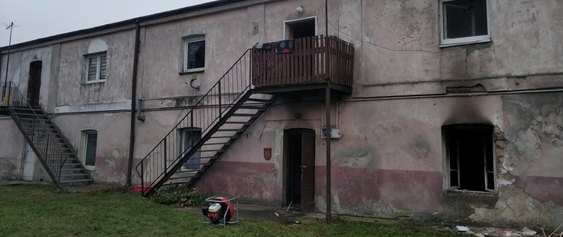 Na zdjęciu widoczny budynek mieszkalny, wielorodzinny, dwukondygnacyjny. Widoczne wypalone okno mieszkania na parterze. Przed budynkiem stoi wentylator, służący do oddymiania.