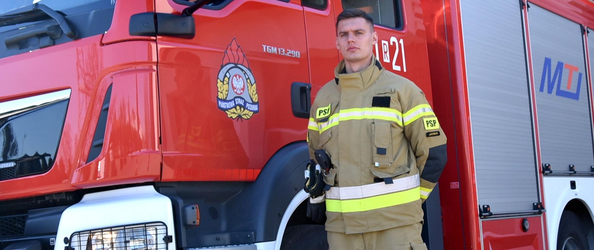 Na zdjęciu strażak w umundurowaniu specjalnym stoi na tle samochodu pożarniczego.