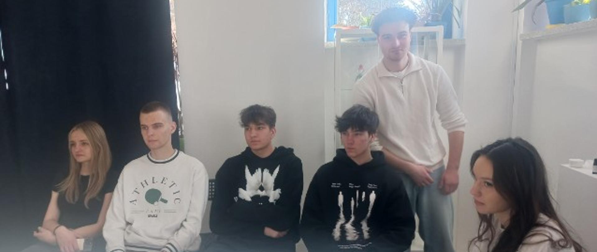 Przy białym stole siedzi trzech młodych mężczyzn i jeden mężczyzna stoi z ich prawej strony. Po obu stronach siedzą dwie młode kobiety. Na stole po prawej stronie leży niebieski długopis i telefon. Za stojącym mężczyzną znajduje się okno a za nim drzewa iglaste. Z tyłu po lewej stronie jest czarna kotara.