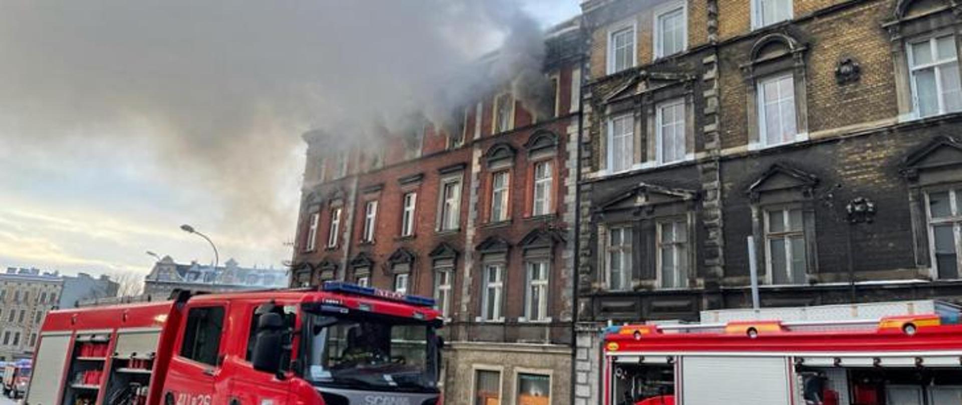 Zdjęcie przedstawia fragment budynku, z którego wydobywają się kłęby dymu. Na pierwszym planie widać dwa samochody gaśnicze.