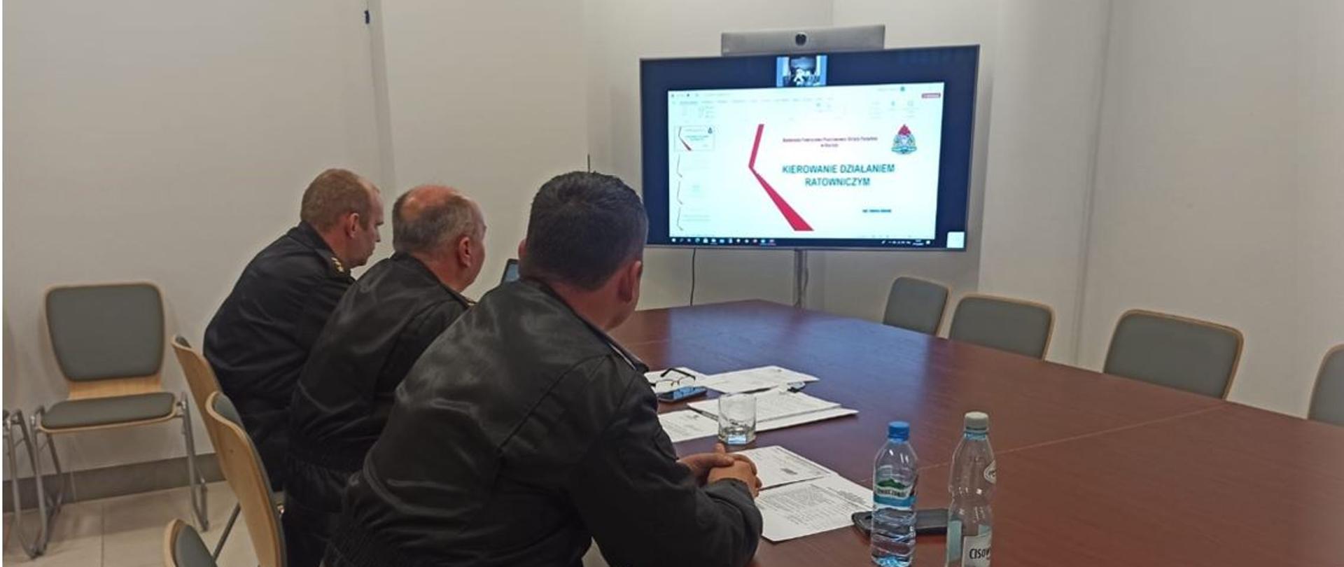 Zdjęcie przedstawia w pierwszym tle strażaków, którzy prowadzą naradę operacyjno-szkoleniową. Siedzą przy stole. W drugim planie widać ekran, na którym wyświetlana jest prezentacja omawiana przez jednego ze strażaków