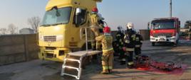 Warsztaty z zakresu ratownictwa technicznego. Złomowisko w Chojnie. Strażacy przystępują do wykonywania dostępu do osoby poszkodowanej znajdującej się w kabinie ciężarówki. Dwóch z nich stoi na podeście ratowniczym, pozostali obserwują działania z ziemi. Za nimi, na macie rozłożony jest sprzęt hydrauliczny: pompa, rozpieracze i nożyce.. W tle wóz straży pożarnej.