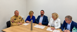 Pięć osób uczestniczy w oficjalnym spotkaniu przy drewnianym stole w sali konferencyjnej. Jedna z uczestniczek podpisuje dokument, pozostali obserwują przebieg podpisania. Na stole leżą przygotowane akty oraz długopis.