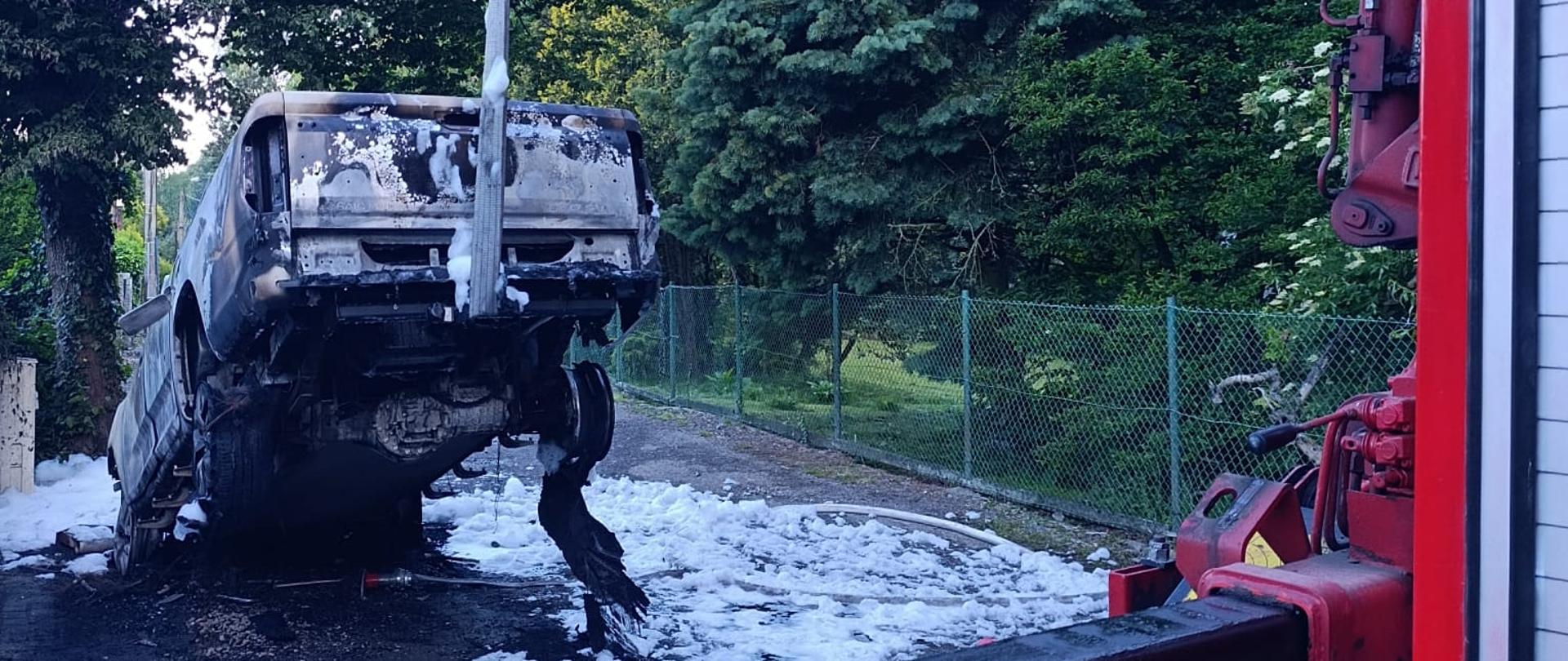 Zdjęcie przedstawia wrak spalonego samochodu podnoszony przez czerwony dźwig hydrauliczny HDS w otoczeniu gaśniczej piany i zielonych drzew.