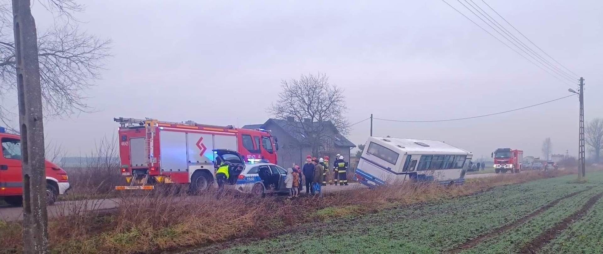 Na zdjęciu widać biało-niebieski autobus, który częściowo znajduje się w rowie a częściowo na poboczu. Widać trzech strażaków. Dwóch w białych hełmach i ciemnych strojach, a jednego w żółtym stroju i czerwonym hełmie. Widać trzy samochody strażackie i jedne policji. Obok radiowozu stoją dzieci, policjant, który wyciąga coś z bagażnika oraz dwie dorosłe osoby. W tle widać dom, drzewa, pole oraz słupy energetyczne. Jest widno.