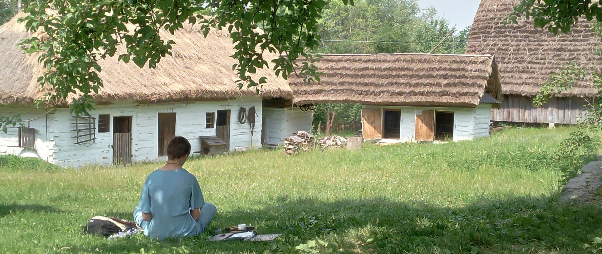 Na pierwszym planie młoda kobieta maluje w cieniu drzewa w tle stare zabudowania wiejskie