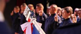 Na pierwszym planie wierzchołek sztandaru Policji. W tle salutujący funkcjonariusze w granatowych mundurach. 