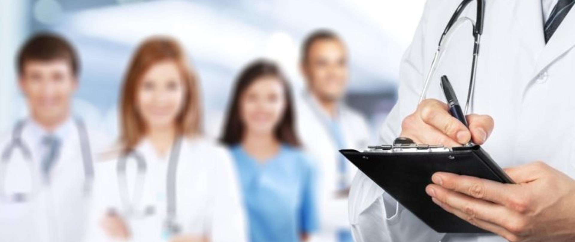 wizerunek lekarza trzymającego podkładkę z dokumentem, w tle inni lekarze