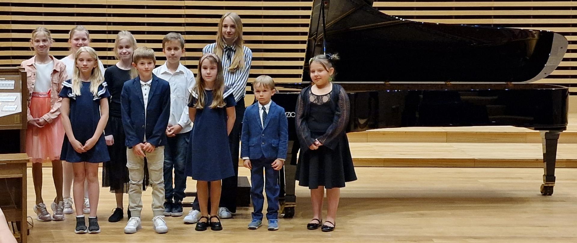  Dziesięcioro dzieci elegancko ubranych stoi przy fortepianie na estradzie sali koncertowej.