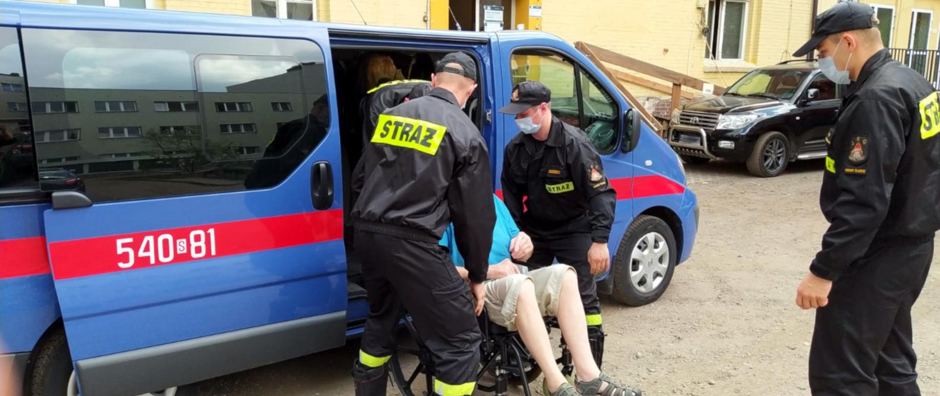 Na zdjęciu widoczna jest ulica przed przychodnią, oznakowany niebieski samochód specjalny Państwowej Straży Pożarnej. Przy samochodzie czterech strażaków w ubraniach koszarowych i maseczkach ochronnych, wynoszą z samochodu mężczyznę na wózku inwalidzkim.