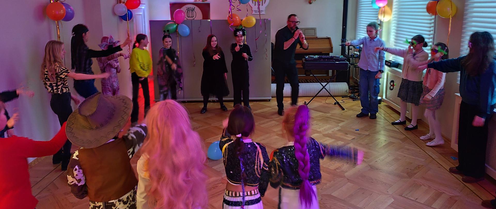 Dzieci w strojach karnawałowych bawiące się w kole w auli szkoły. Sala ozdobiona balonami.