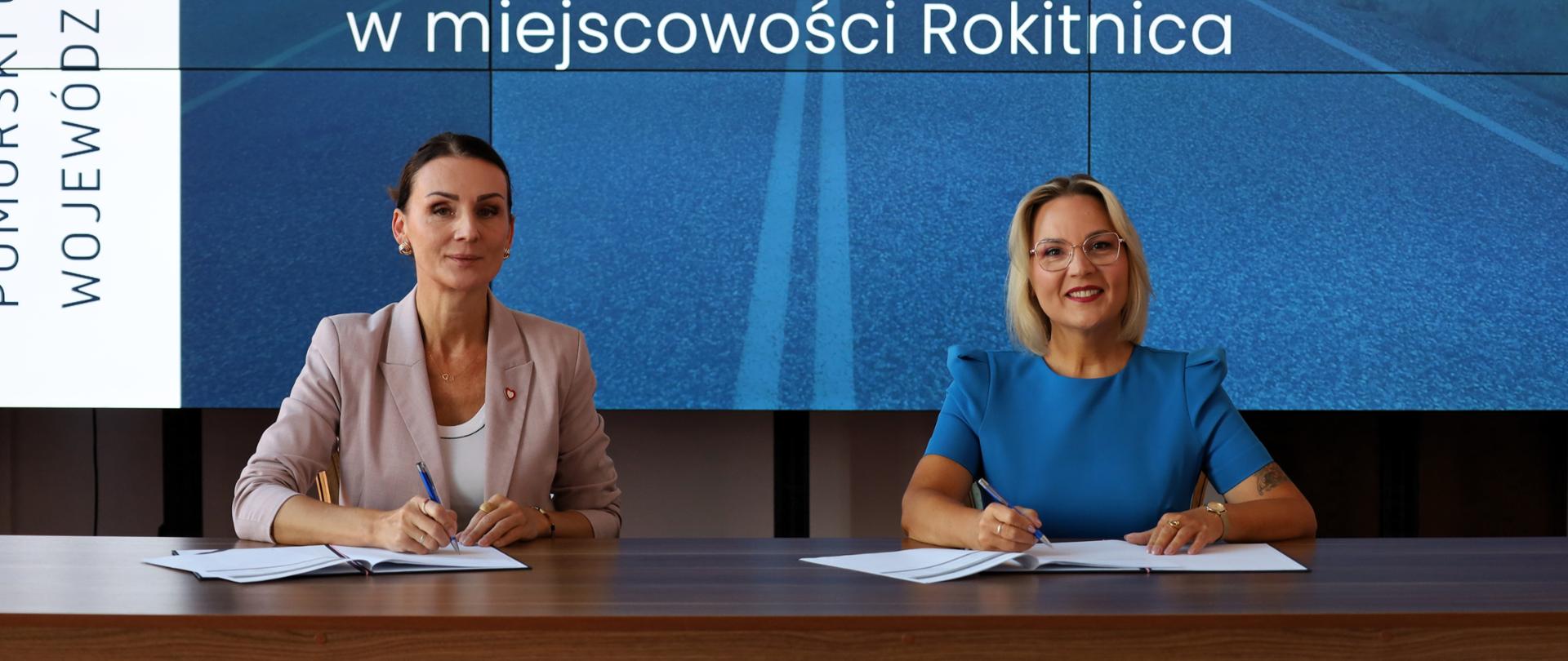 Dwie kobiety siedzą przy drewnianym stole i podpisują dokumenty, a za nimi znajduje się duży ekran wyświetlający informacje o dofinansowaniu budowy dróg gminnych w miejscowości Rokitnica w gminie Pruszcz Gdański na kwotę 2 879 428,60 zł.