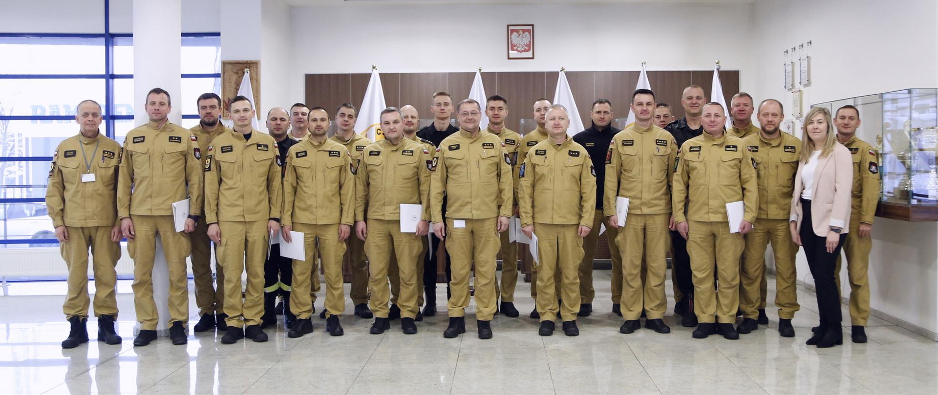 Grupowe zdjęcie funkcjonariuszy PSP (19 osób) w towarzystwie Zastępcy Komendanta Centralnej Szkoły PSP, kobiety oraz trzech funkcjonariuszy CS PSP