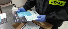 Zdjęcie przedstawia część tułowia strażaka OSP trzymającego w rękach ulotkę propagujące wiedzę na temat profilaktyki oraz szczepień przeciwko COVID-19.