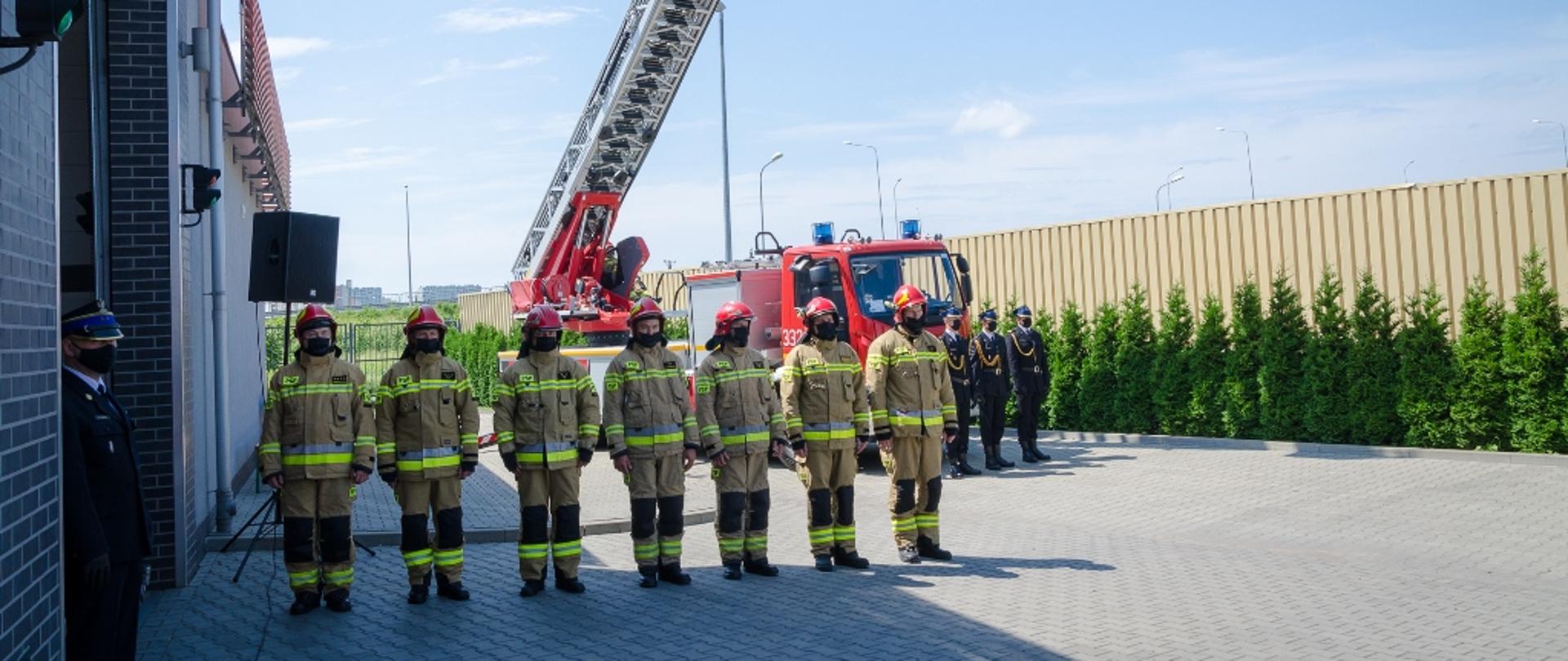 Wręczenie promes mazowieckim OSP – na zdjęciu siedmiu strażaków. Na drugim planie samochód strażacki z drabiną, na której powiewa flaga biało-czerwona.