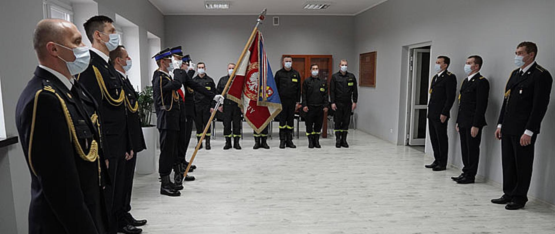 strażacy stoją w pozycji zasadniczej słuchając mazurka Dąbrowskiego, po lewo trzech strażaków ubranych w mundur galowy ze sznurem w maseczkach ochronnych na twarzy, po środku na lewej stronie sali trzech strażaków ubranych w mundur galowy ze sznurem w maseczkach ochronnych na twarzy stoi w poczcie sztandarowym, jeden trzyma sztandar a dwóch oddaje honor podczas śpiewu hymnu państwowego, w głębi sześciu strażaków ubranych w umundurowanie koszarowe z maseczka ochronną na twarzy stoi w pozycji zasadniczej, po prawej stronie zdjęcia stoi trzech strażaków w mundur galowy ze sznurem w maseczkach ochronnych na twarzy w pozycji zasadniczej
