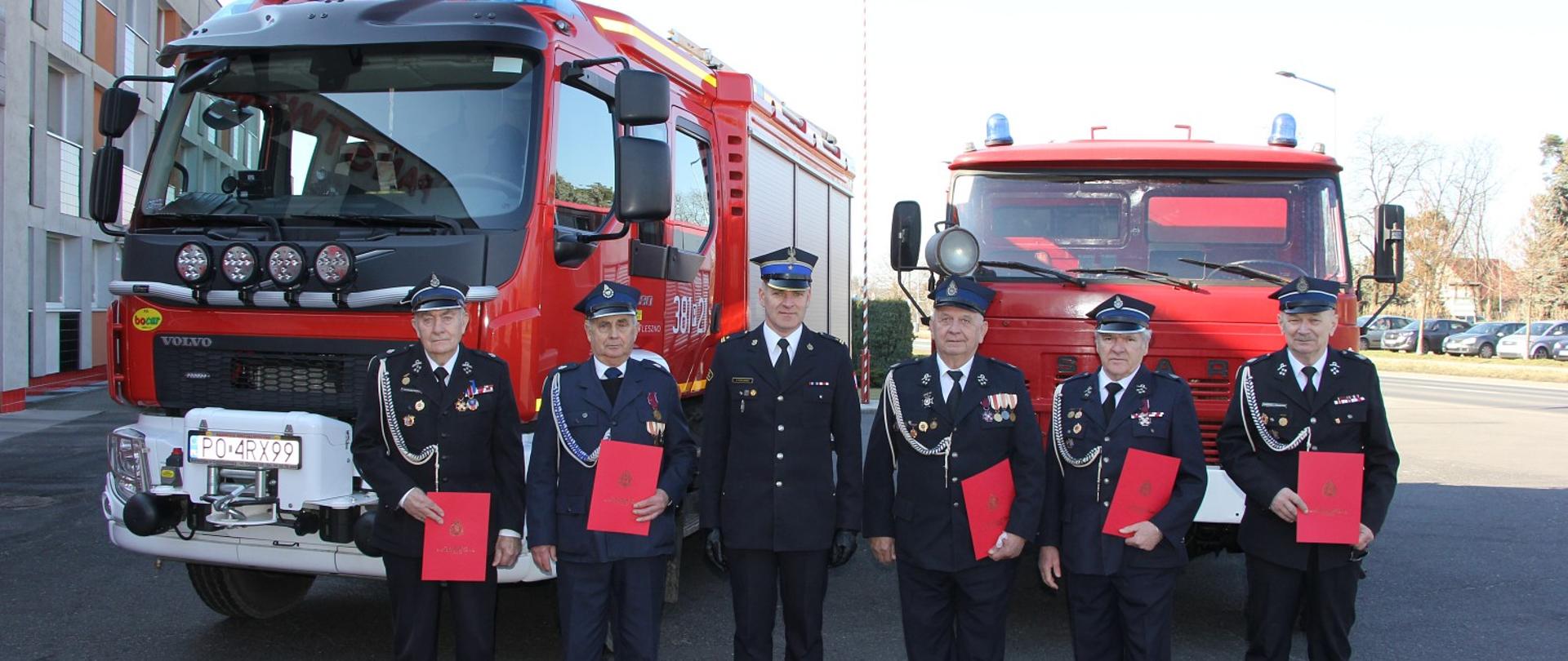 strażacy OSP stojaa w szeregu w mundurach wyjściowych, w środku stoi strażak PSP, za nimi dwa czerwone strażackie samochody