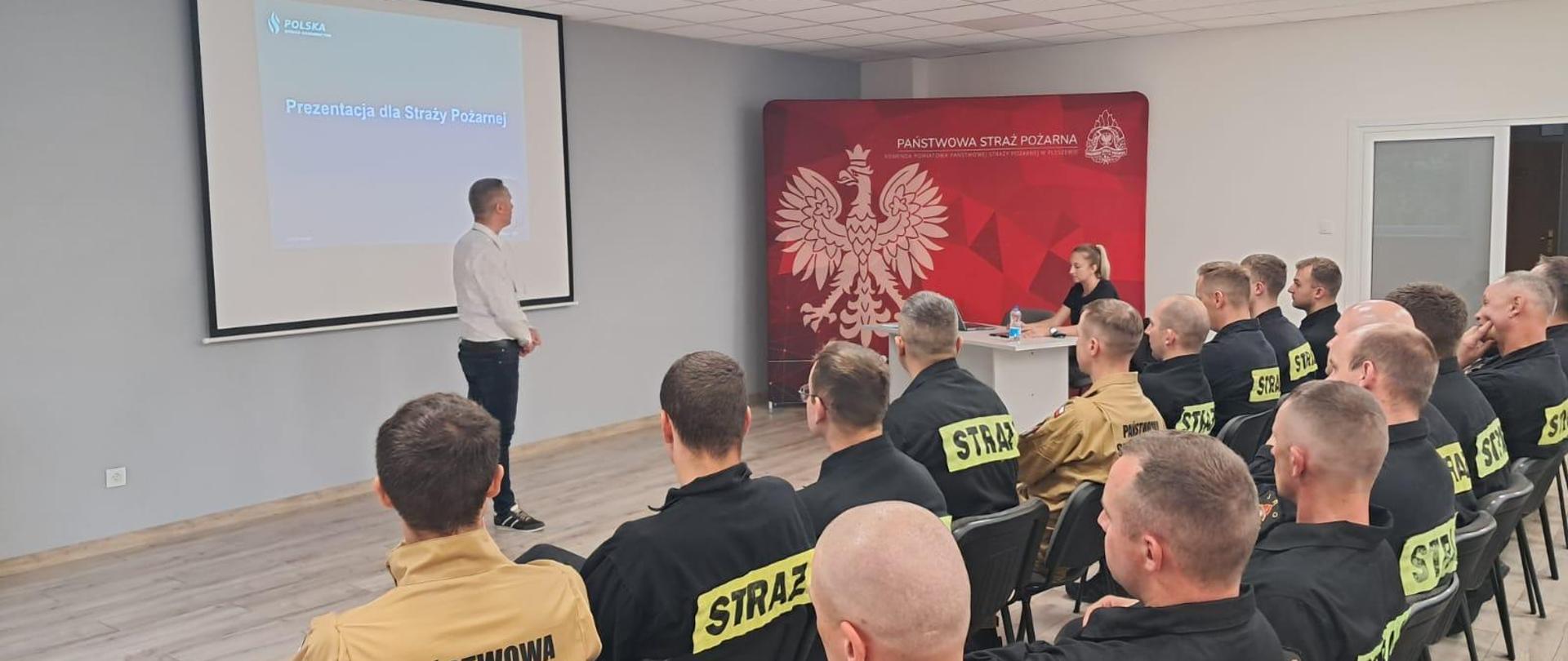Na oświetlonej sali siedzi grupa mężczyzn ubranych w mundury straży pożarnej, przed nimi przemawia mężczyzna w stroju cywilnym, za nim widoczny jest ekran i strona prezentacji medialnej, obok widoczna siedziąca kobieta z laptopem za która stoi baner Państwowej Straży Pożarnej