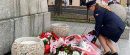 79 rocznica wybuchu Powstania Warszawskiego. Przed pomnikiem Żołnierza Polskiego policjantka składa wiązankę biało-czerwonych kwiatów. W tle pozostałe delegacje, drzewa i zabudowania.