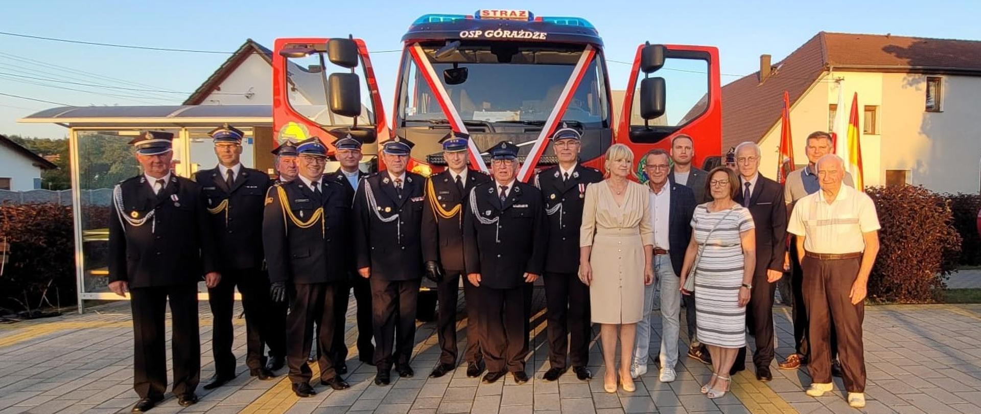 Na tle przodu samochodu pożarniczego z otwartymi drzwiami stoją najważniejsi goście zaproszeni na uroczystość przekazania. Przedstawiciele PSP i OSP, organów administracji rządowej i samorządowej oraz zarząd i zasłużeni działacze OSP Górażdże.