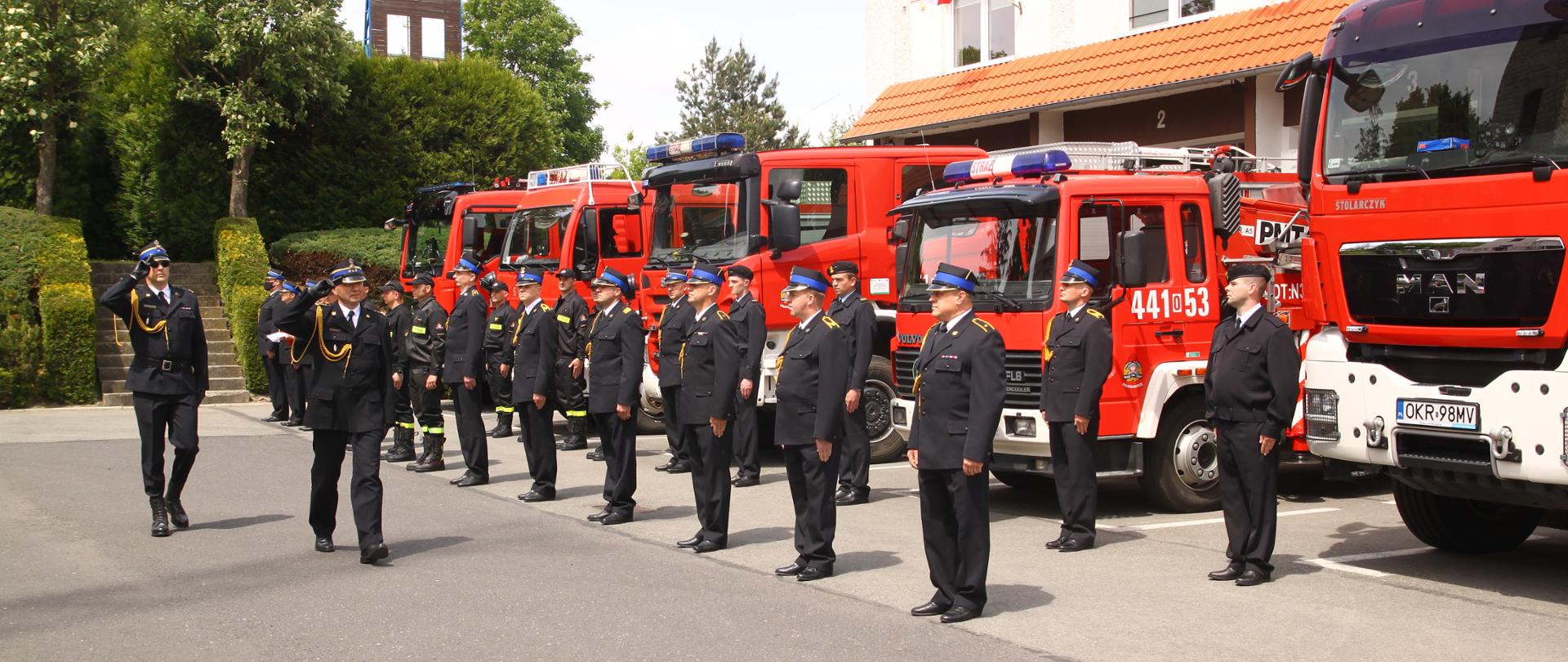 Z-ca Opolskiego Komendanta Wojewódzkiego PSP w asyście dowódcy uroczystości dokonuje przeglądu pododdziału strażaków z KP PSP Krapkowice. W tle samochody pożarnicze i budynek JRG. Strażacy w umundurowaniu galowym.