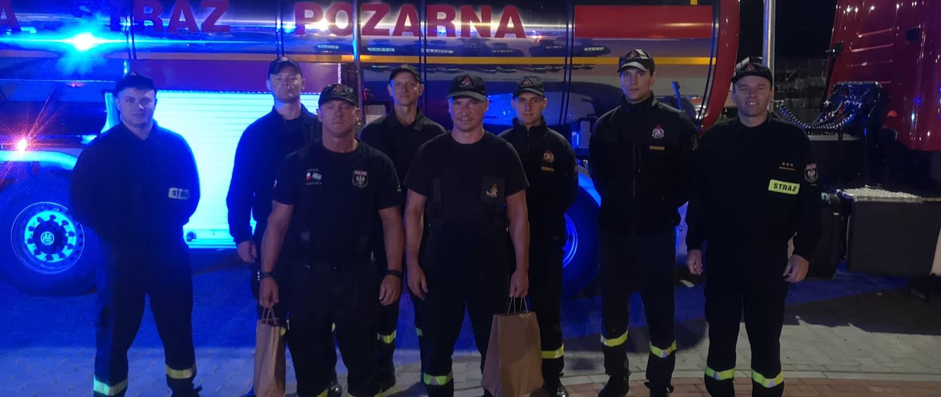 Zdjęcie przedstawia strazaków stojących przed autocysterną strażacka z włączonymi światłami niebieskimi błyskowymi.