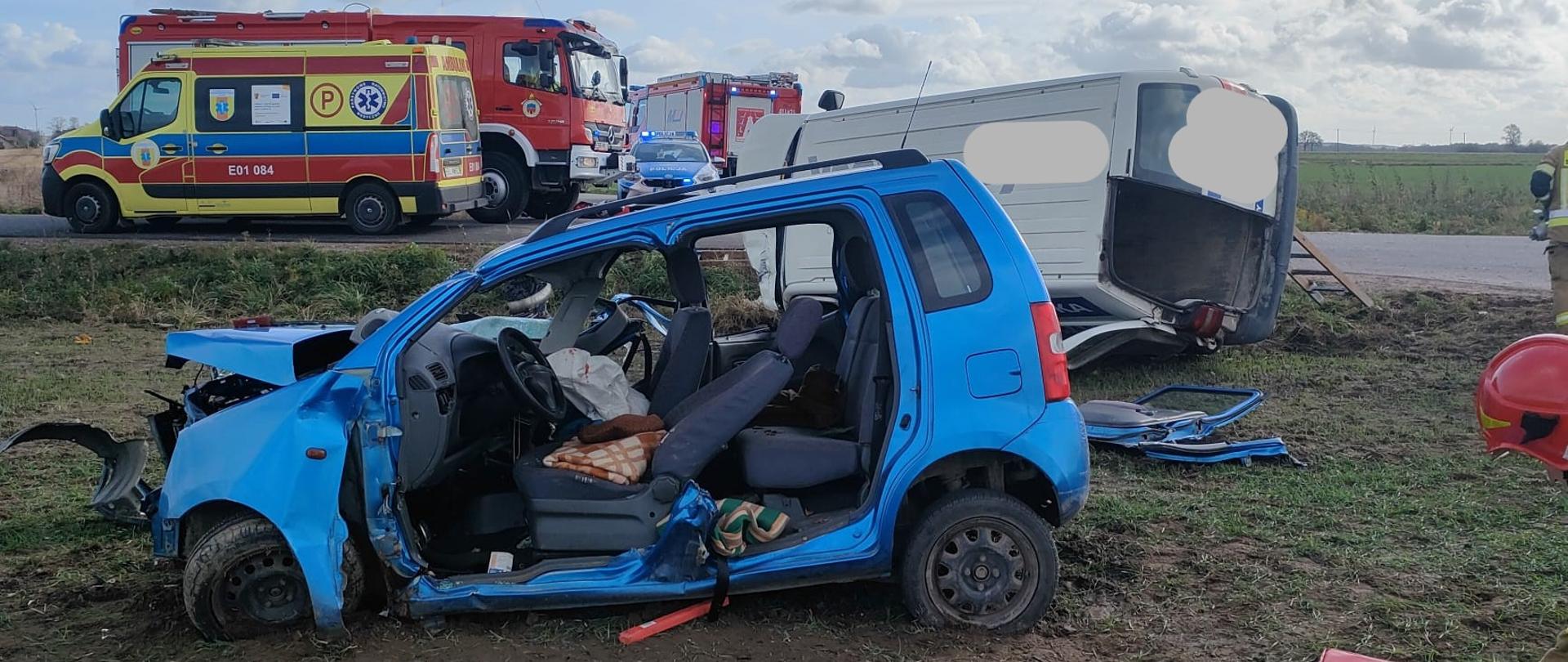 Niebieski samochód osobowy, który brał udział w wypadku, uszkodzony cały przód pojazdu, strażacy przy użyciu narzędzi hydraulicznych wycieli przednie i tylne drzwi pojazdu, fotel pasażera całkowicie rozłożony do tyłu, wystrzelona poduszka kierownicy, przy pojeździe rozłożona mata sprzętowa, na której leży torba medyczna, za samochodem widać samochód dostawczy który uczestniczył w wypadku, leży na lewym boku, ma wyrwane jedne drzwi z tyły pojazdu. W oddali za pojazdami widać ambulans, radiowóz policyjny i dwa wozy strażackie