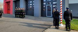 Trzech strażaków stoi obok masztu flagowego i jeden ją przypina. Pozostali strażacy stoją w dwuszeregu. W tle zdjęcia budynek garażu Komendy z napisem Państwowa Straż Pożarna.