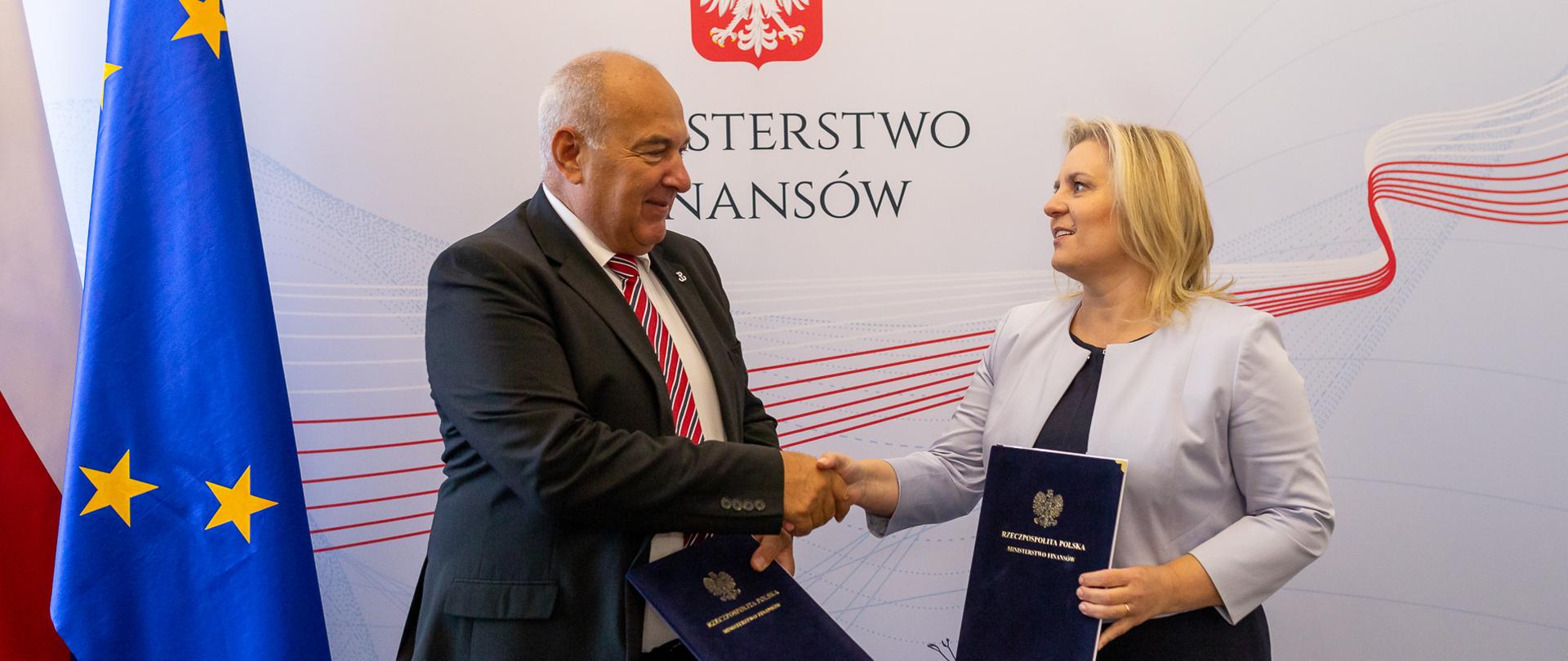 Uścisk dłoni po podpisaniu porozumienia - minister finansów Tadeusz Kościński i kierująca Polską Agencją Inwestycji i Handlu Grażyna Ciurzyńska na tle baneru MF i flag Polski i UE